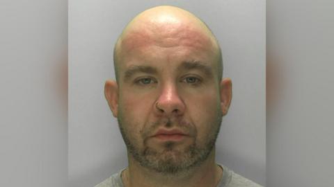 A mugshot of Kane Kotwica, a bald man with stubble on his face and a nose ring. He has a blank expression as he stares directly at the camera against a light grey background, and is wearing a light grey T-shirt.