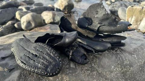 Old black leather shoes, which are soaking wet and partially ruined, sat on a sandy beach with rocks behind.