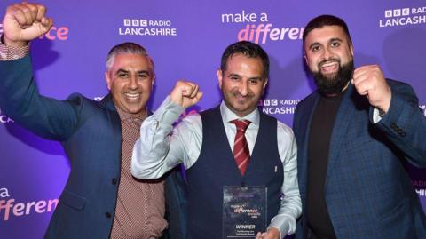 Three men from Whitefield Youth Association, dressed smartly, smiling and raising their fists in triumph