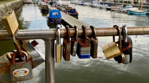 A close-in view of padlocks attached to the metalwork of Pero's Bridge in the centre of Bristol. Behind the waterways of the floating harbour are visible along with some houseboats