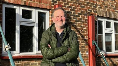 A man with short grey hair wearing a green coat looking into the camera with his arms folded. Behind him are terraced houses propped up with metal bars