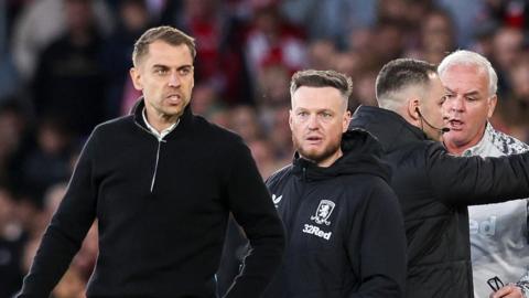 Tonda Eckert has a stern grimace on his face as Kim Hellberg looks on while the fourth official speaks to Middlesbrough coach Adi Viveash behind Hellberg