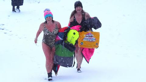Two women in bathing suits - one leopard print, one black - walking along a snow covered beach. They are wearing woolly hats and thermal boots but have bare legs and arms. They're carrying bags with colourful swimming kit and have bright smiles on their faces.