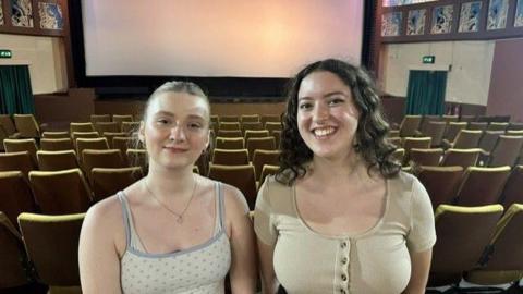 Two women in pale coloured tops, one with blonde hair and other with brown hair, standing in a cinema with rows of seats and a large screen behind them