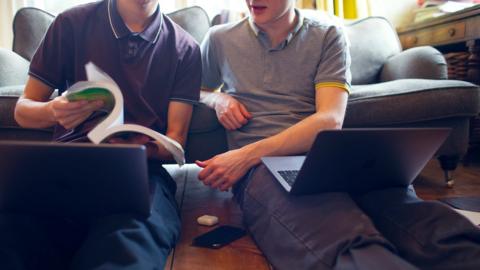 Two GCSE maths students share a past papers textbook while looking at their own laptops in front of the sofa.