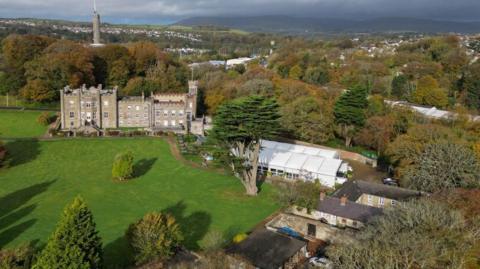 The Nunnery from above. The site comprises of large green grassy areas, an old grey building with turrets. A white marquee has been erected on the grounds. Lots of trees and houses can been seen in the background.