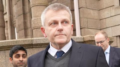 Deputy Philip Ozouf is outside Jersey's Royal Court. He's wearing a suit and is looking away from the camera. He is being followed by two men in suits, one with a microphone.
