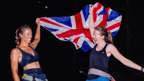 The picture shows two rowers celebrating on the deck of a boat at night. One of them is holding a large Union Jack above her head, letting it billow out behind her, while the other stands beside her with an arm raised in triumph. Both look exhausted but jubilant, as if they have just completed a major challenge at sea.