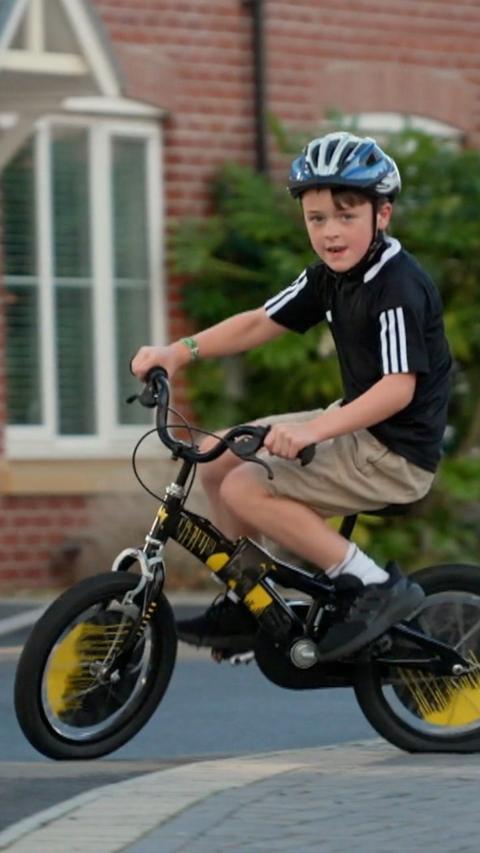Rex is riding a bicycle. He has a blue helmet on and wears a black top with white stripe detail on the sleeves and beige chino shorts with black trainers.