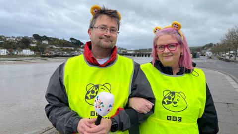 Michael Chequer is seen arm in arm with Beki Sharples. They are both wearing high-visibility vests with the Children in Need logo and fluffy ears on their heads. They are standing on a waterfront.