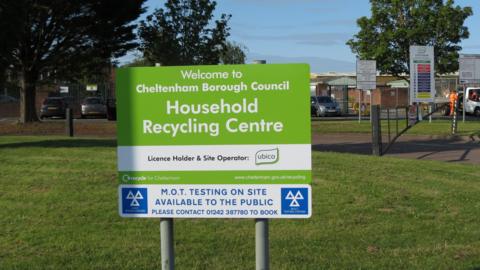 A green sign for Swindon Road Recycling Centre in Cheltenham that reads "Welcome to Cheltenham Borough Council Household Recycling Centre". The sign is on a grass verge at the front of the site. The recycling centre can be seen in the background along with a car park and men in orange hi-vis clothing in a van.