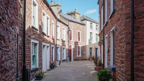 The view of an alleyway between two rows row of quaint red brick cottages. They have brightly coloured window frames and the sky is blue.