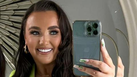 Chloe Ferris smiling at the camera while taking a selfie.  She has long, wavy dark hair and long, painted fingernails.  She wearing make-up, earrings and a lime green outfit. 