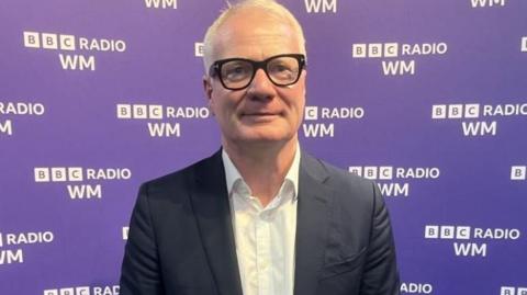 Richard Parker in black jacket, white shirt and glasses, standing against a purple background with the 鶹ҳ Radio WM logo across it