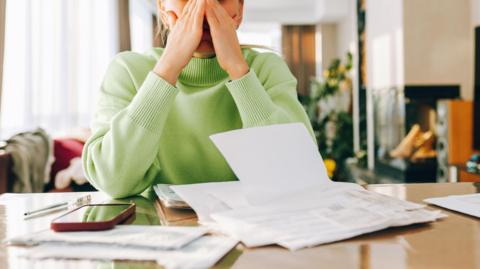 A stock image picture of a woman in a light green jumper with letters and a mobile phone in front of her. She has her hands up to her face, looking tired or stressed.
