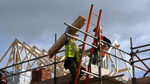 A stock image of builders working on a roof. One is up a ladder in a high visibility vest while holding some timbers.
