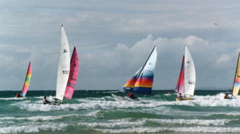 A series of Hobie Cats sail in green surf and waves. The sky is cloudy behind them. 