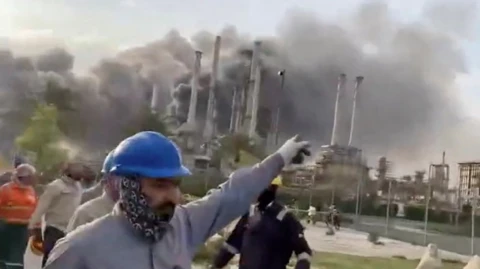 Workers outside oil refinery in Saudi Arabia with smoke billowing after drone attack
