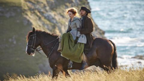 Ross and Demelza Poldark are sitting on a chestnut horse which is walking through long grasses on a sunny day. Behind them is a sheer cliff and sparkling sea. Demelza is riding side saddle and is wearing a long dress, cloak and gloves while Ross is wearing a tricorn hat, heavy overcoat and gloves. 