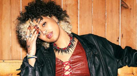 Nxdia with blond and black hair rests her head to the side on her hand. She is wearing a red leather-look top with a leather jacket and black and red choker necklace with spikes.