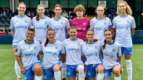 A women's football team posing on a pitch. The players are wearing a white and blue uniform, except for the goalkeeper who is wearing a pink, purple and white uniform.