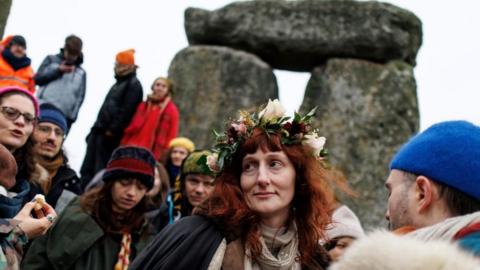 A woman with red hair wearing a flower crown stands among a crowd of people in front of some of the stones that makeup Stonehenge.