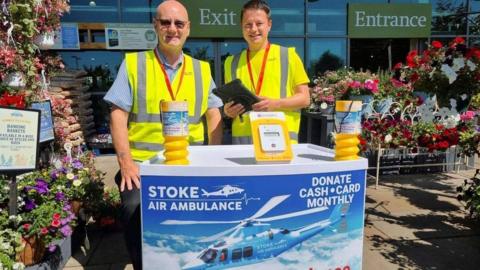 Two men in high-vis jackets standing behind a counter advertising plans for Stoke Air Ambulance, outside a garden centre