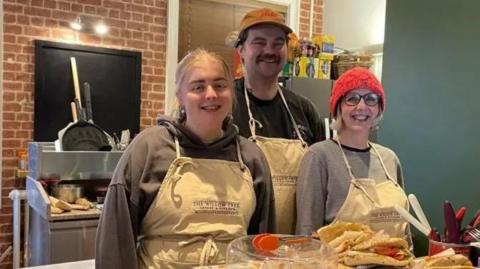 Volunteers Ellie and Jack standing next to Jo Gordon behind the till at the Willow Tree Coffee and Kitchen. Pastries and sandwiches are displayed on the counter in front of them. They are all wearing white aprons. Ellie has straight blonde hair tied in a ponytail. Jack is wearing a black cap and has moustache. Jo has short, straight grey hair and she is wearing round glasses and a red beanie hat.