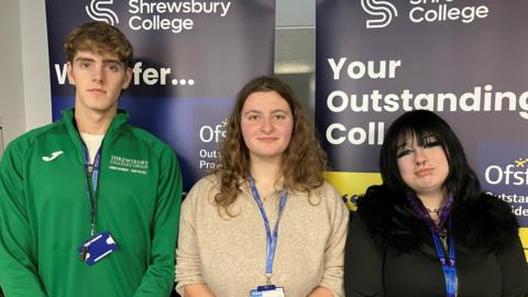 A tall youth with blonde curly hair and a green hoodie next to a young woman with long blonde wavy hair and a beige jumper and a shorter young woman with black hair and a black top