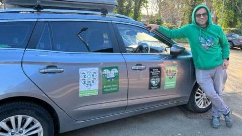 Sâmân Jamshidifard smiling for the camera while leaning on his car. He is wearing a green hoodie. He is outside on a sunny day, another car can be seen behind him.