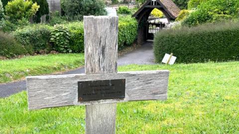 A wooden cross in the ground, with a plaque on it. Behind it is a hedge and a covered gateway into the graveyard.