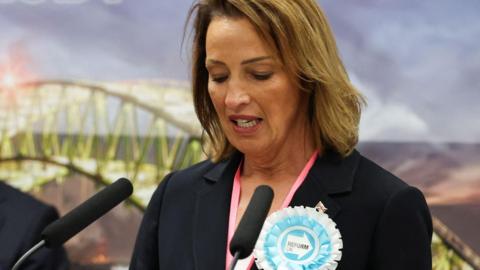 Sarah Pochin has medium length brown hair, a blue jacket and a large sky blue Reform rosette. A photo of the Runcorn Bridge is in the background.