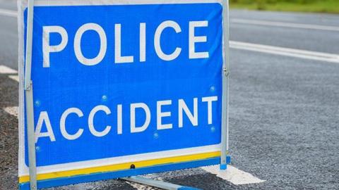 A sign saying POLICE ACCIDENT in white letters, on a blue background. It is on a road bordered by greenery.
