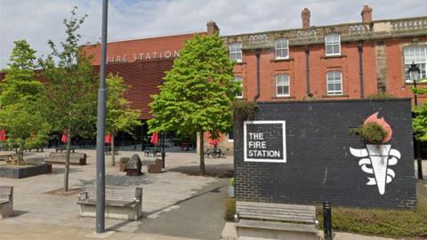 Brown building in background with signage that says "Fire Station" with a black box in front which says "The Fire Station" and an image of a torch with fire. 
