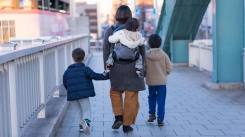 A stock image of a woman walking with three children of differing ages. The photograph is taken from behind.