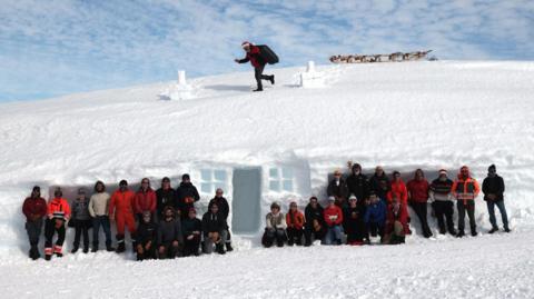 Group of around 30 people in front of a snow house with someone dressed as Santa on the roof.