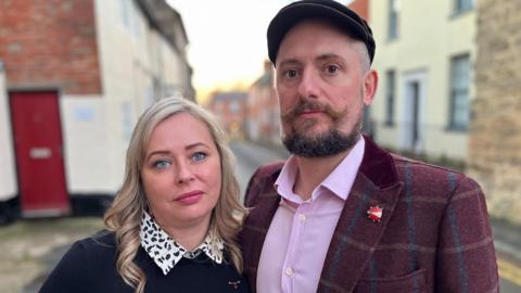 Couple Carly and Carl are pictured standing together in the street. There are terraced houses in the background. Carly has blonde hair and is wearing a black and white shirt and a dark jumper. Carl is wearing a cap and has a beard and moustache. He is wearing a lilac shirt with a maroon checked jacket. He has a ladybird brooch on the lapel