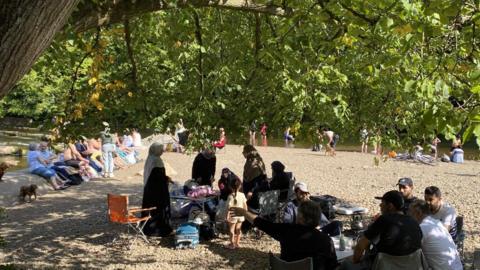 Groups of people sit on a river bank and paddle in the water. It is a sunny day and the site is shielded by trees. 