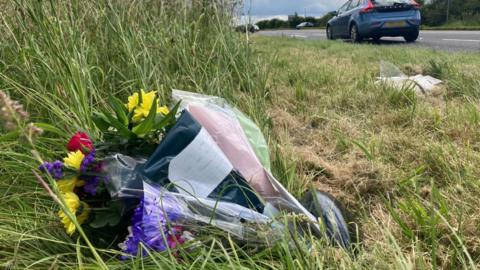 Flowers by the side of Newark Road, North Rauceby, where two teenager suffered fatal injuries in a car crash. There are multiple bunches wrapped in cellophone placed in long grass. In the top right corner of the photo you can see a car driving along the road.