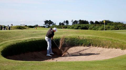 A wide shot of a golfer wearing a dark polo shirt, light grey trousers and a white cap chipping a golf ball out of a bunker. Sand can be seen spraying up where his club and the ball made contact and the ball has lifted just out of the bunker. In the distance the yellow flag marking the hole can be seen as well as some scrubland and, further up the image, the blue sky.
