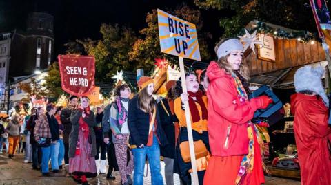 A large group of people dressed in orange clothes walking past a row of Christmas market huts at night. Two people are holding up signs - one saying "rise up" and the other saying "seen & heard".