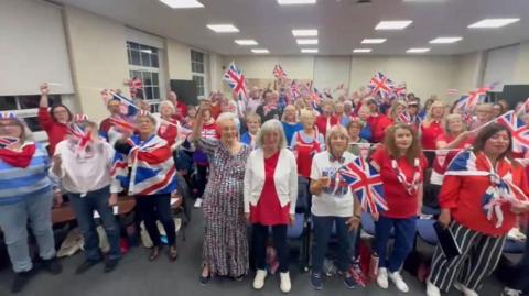A choir stands in a group looking at the camera and waving union jack flags and many wearing the same blue white and red colours.