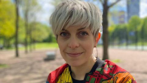 Head and shoulders shot of Linn who has short light blonde hair and is wearing a colourful red, yellow and orange patterned bomber jacket.