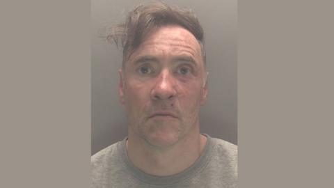 A mugshot of Paul Doyle wearing a grew jumper looking stunned. His hair, a long brown fringe, is dishevelled and to the side. He is wearing a grey t-shirt.