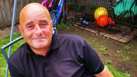 The late Joseph Tidmarsh, who was bald and had a slight stubble. He is photographed wearing a navy polo top. He is sitting in a garden with bikes and balls in the background. He is smiling.