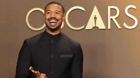 Michael B Jordan - a black man with short, dark hair, a moustache and beard - grins widely while clutching a golden Academy Award. He is wearing a dark buttoned jacket and is stood in front of a wall with the words "Oscars" printed on it in large capital letters.