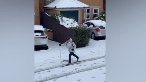 A man skiing down a residential street. He is wearing a grey jacket and black trousers.