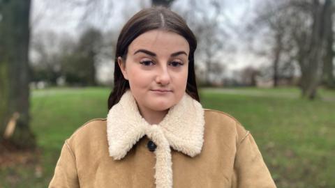 A woman with long brown hair, wearing a brown jacket with woolly collar, looks at the camera.