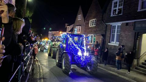 Tractors are driving through a village street strung with fairy lights They are blue and green and there are people lining the streets either side watching them pass