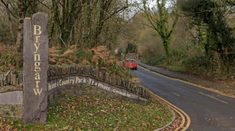 A tree-lined country road curves gently through a wooded landscape at the entrance to Bryngarw House and Country Park. In the foreground, a tall vertical stone marker carved with the word “Bryngarw” stands beside a curved stone wall bearing bilingual Welsh and English signage for the park. Brown ferns, ivy-covered trees and fallen leaves surround the entrance. A small red car is visible further down the road, which disappears into the wooded park beneath a canopy of bare branches.
